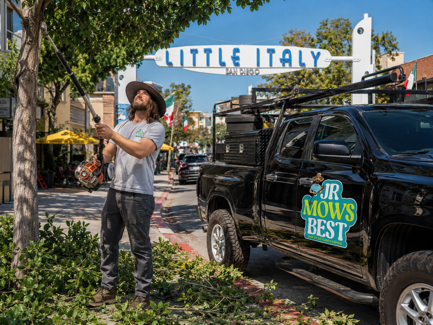 Jr trimming a street tree beside his Jr Mows Best truck under the Little Italy sign in downtown San Diego